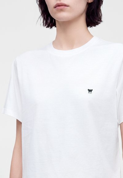 Femme aux cheveux courts et foncés portant un t-shirt blanc uni avec un petit logo papillon noir sur le côté gauche de la poitrine.