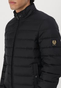 Čierna prešívaná bunda s vysokým golierom, so zipsom a nášivkou loga Belstaff na ľavom rukáve.