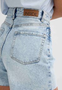 Bermudas de mezclilla de color azul claro con cintura alta, con una etiqueta de cuero marrón que dice "INSIDE" y dos bolsillos traseros con costuras en contraste.