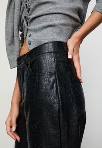 Nærbillede af person iført højtaljede sorte faux krokodillelæderbukser og en grå kort, knaplukket sweater, hånd hvilende på hoften.