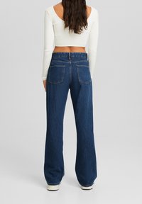 Jeans in denim a vita alta e gamba larga di colore blu scuro. Presentano due tasche posteriori e una chiusura con zip. Il materiale appare resistente con una texture liscia.