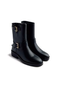 Botines de cuero negro hasta el tobillo con punta redonda, acentos de hebilla dorada y suela de goma plana. Presenta tirantes laterales y un acabado liso.