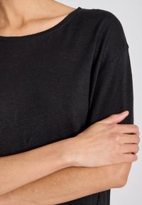 Osoba v černém tričku s krátkým rukávem má jednu ruku složenou přes tělo a druhou ruku spočívá na opačném horním rameni.