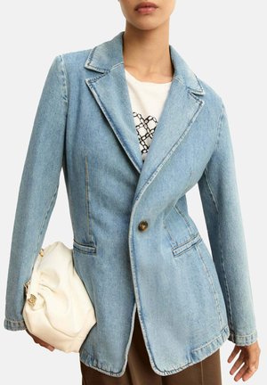 Vrouw draagt lichtblauwe denim blazer over wit overhemd, houdt zachte witte handtas vast, met bruine broek tegen een effen achtergrond.