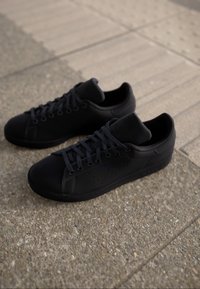 Černé tenisky s hladkým syntetickým svrškem, kulatou špičkou, perforovanými detaily a plochými tkaničkami. Vyznačují se polstrovaným jazykem a gumovou podrážkou.