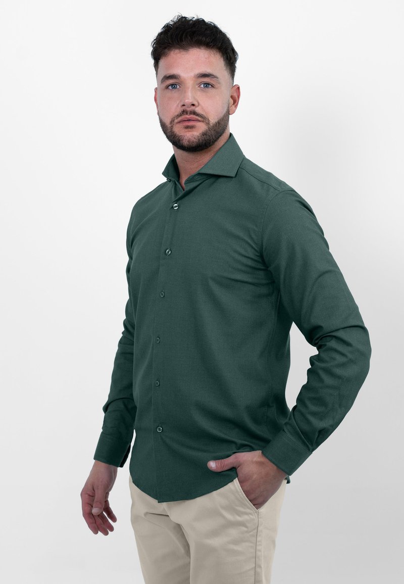 Donkergroene langemouwen shirt met een knoopsluiting aan de voorkant, kraag en textuurstof. Draag het met beige broeken, handen in de zakken.