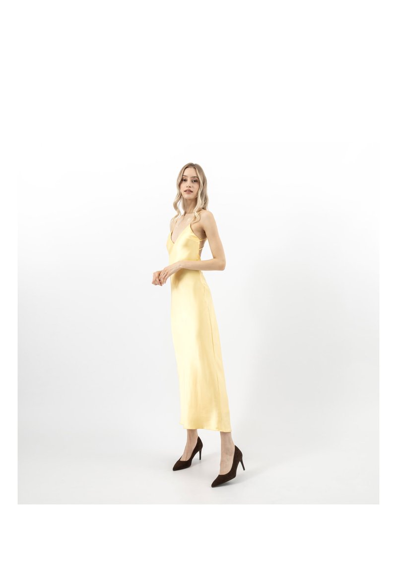 Donna in piedi di profilo, indossa un abito sottoveste in satin giallo e tacchi alti neri su uno sfondo bianco.