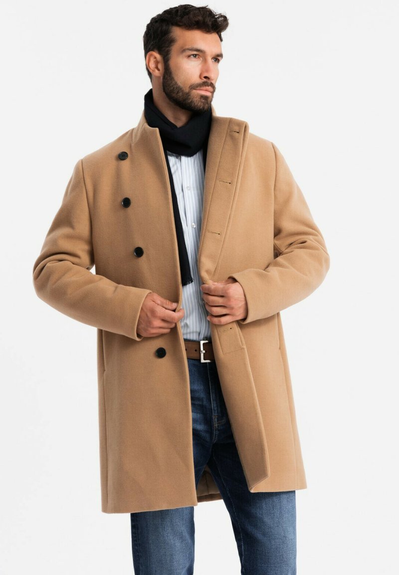 Abrigo de mezcla de lana en color beige con un diseño de doble botonadura, que presenta botones negros y un bolsillo tipo solapa en la parte delantera. Llevado sobre una camisa a rayas y unos jeans.