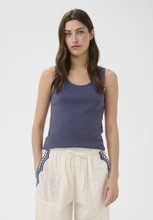 Femme aux longs cheveux bruns portant un haut sans manches bleu foncé et un pantalon blanc à cordon avec bordure en dentelle bleue sur les poches.