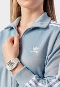Šviesiai mėlynas zip-up džemperis su baltu Adidas logo, derinamas su baltu skaitmeniniu laikrodžiu, turinčiu stačiakampį ciferblatą ir tekstūruotą dirželį, rodančiu laiką.