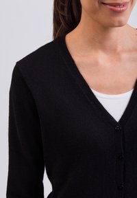 Svart kofta i mjukt stickat material, med V-ringning, lång ärm och sex svarta knappar på framsidan för stängning.