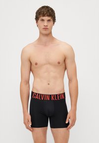 Boxeri negri cu logo-ul "CALVIN KLEIN" în roșu pe banda elastică, design ajustat, textură moale și lungime până la mijlocul coapsei.