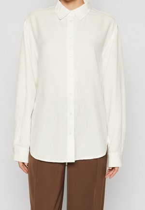 Person iført en ensfarvet hvid skjorte med lange ærmer og brune bukser, stående foran en neutral baggrund.