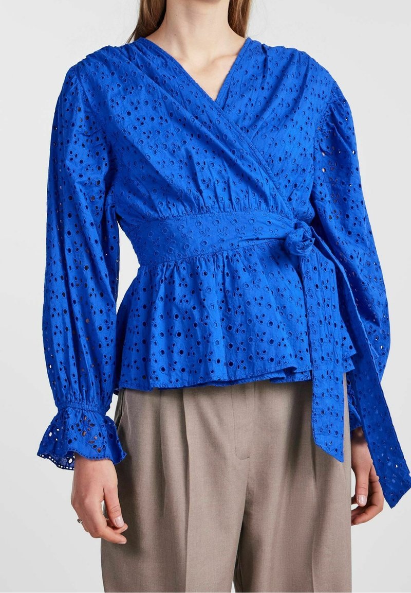 Femme portant un chemisier cache-cœur bleu en broderie anglaise avec de longues manches bouffantes et des poignets à volants, associé à un pantalon beige à jambes larges.