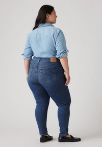 Calças de ganga em lavagem escura com cintura alta, corte slim e bolsos traseiros. Combinadas com uma camisa de botão em chambray azul claro.