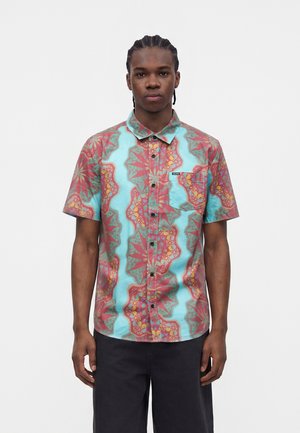 Junger Mann mit geflochtenem Haar, der ein kurzärmliges Hemd mit türkisfarbenen und roten geometrischen Blumenmustern und schwarze Hose trägt.
