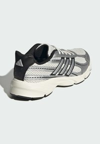 Šedá a biela športová obuv s vrchom z výpletového materiálu, čiernymi akcentmi, reflexnými pruhmi a polstrovanou podrážkou. Má systém šnurovania a textúrované detaily.