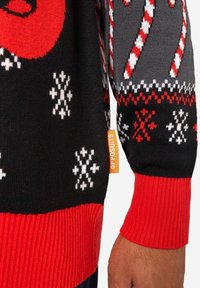 Crni pleteni džemper s uzorcima crvenih, sivih i bijelih štapića i pahuljica. Uključuje crveni rebrasti rub i narančastu oznaku marke.