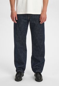 Jeans in denim blu scuro con vestibilità rilassata, caratterizzati da cuciture a vista e indossati con scarpe nere. La trama è liscia con una leggera lucentezza.