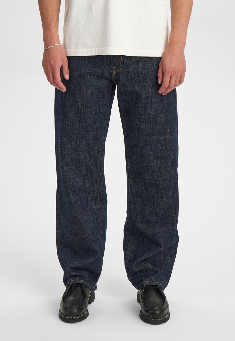 Jeans in denim blu scuro con vestibilità rilassata, caratterizzati da cuciture a vista e indossati con scarpe nere. La trama è liscia con una leggera lucentezza.