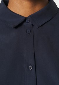 Námořnická modrá košile s hladkým povrchem, s klasickým límcem a dvěma knoflíky na předním zapínání.