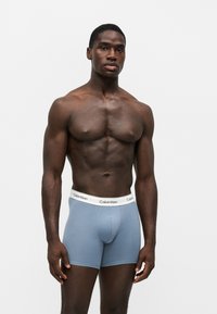 Homme musclé debout sur fond blanc, portant un boxer Calvin Klein bleu clair avec une ceinture blanche.