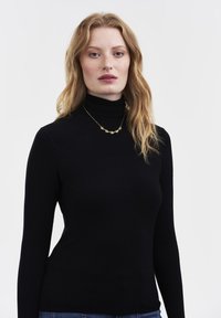 Noor naine pikkade punaste juustega, kellel on seljas must kõrgekaelusega pluus ja õrn kuldne lilleline kaelakee, seisab ühevärvilise valge tausta ees.