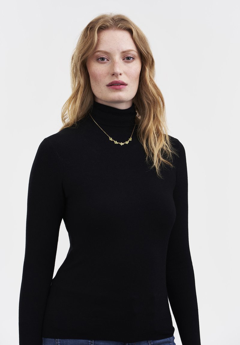 Noor naine pikkade punaste juustega, kellel on seljas must kõrgekaelusega pluus ja õrn kuldne lilleline kaelakee, seisab ühevärvilise valge tausta ees.