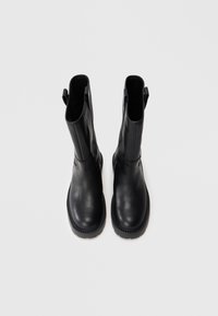 Botas de tobillo de cuero negro con una textura suave, puntera redondeada, dos lengüetas en la parte superior y suela de goma gruesa, vistas desde arriba.