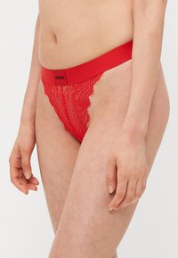 String rouge en dentelle avec une ceinture élastique rouge contrastante arborant le nom de la marque "HUGO". La dentelle présente un motif à bord festonné.