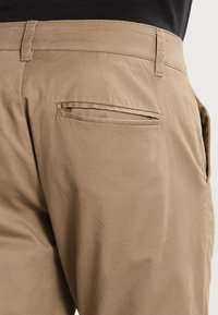 Zbliżenie na beżowe spodnie ukazujące tylną kieszeń z patką i szlufki na paski u osoby noszącej czarną bluzkę.