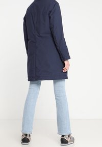 Mörkblå vadderad kappa, knälång, med rak passform. Kombinerad med ljusblå utsvängda jeans och svarta sneakers. Slät tygtextur.