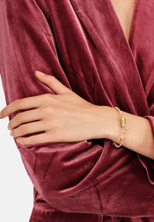 Bracciale color oro con perline decorative, con una chiusura liscia e ovale. Indossato su una mano contro un tessuto di velluto bordeaux strutturato.