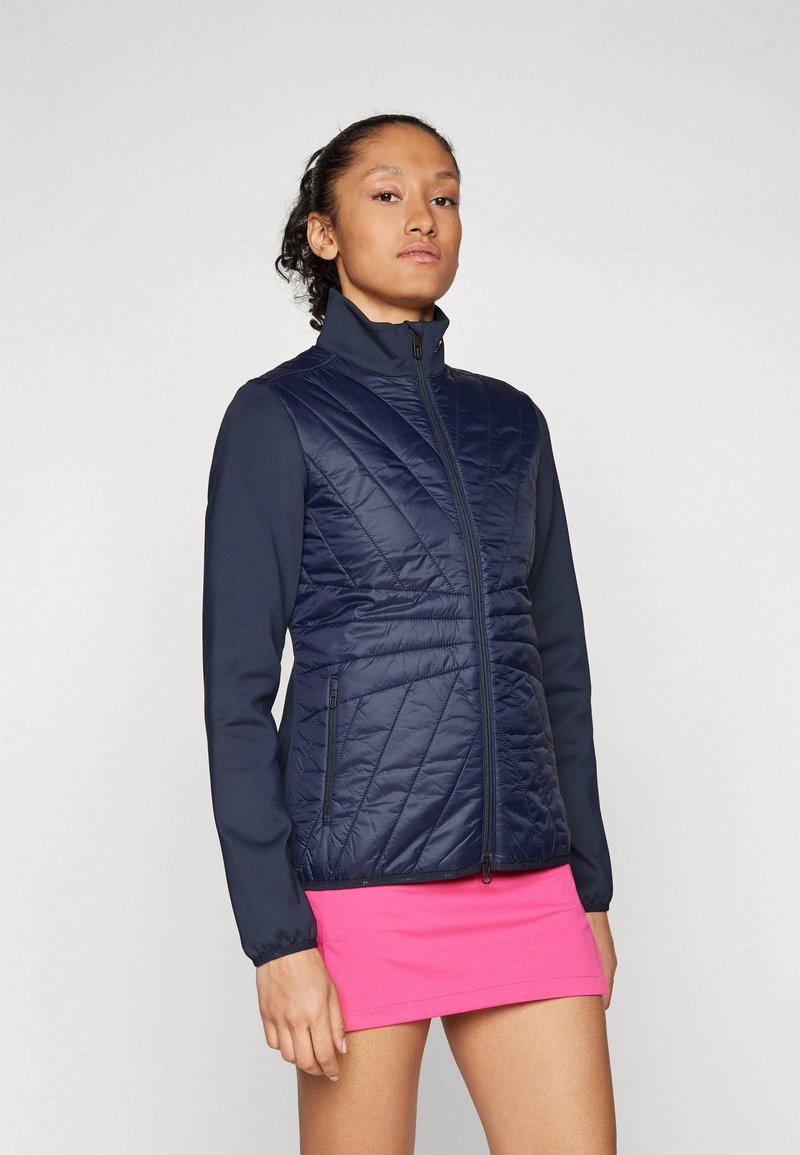 Marinblå quiltad jacka med hög krage och långa ärmar; har en dragkedja och sidofickor; bärs över en rosa kjol.