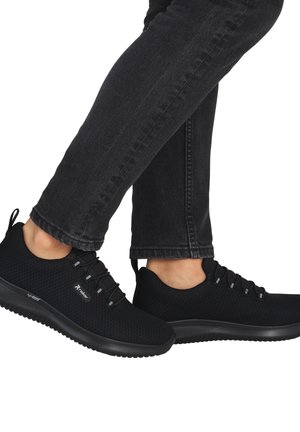 Person trägt schwarze Netz-Sneakers mit elastischen Schnürsenkeln und schwarze Jeans, geht mit einem leicht angehobenen Fuß.