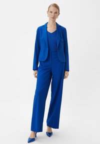 Kobieta stoi w królewskich niebieskich garniturze z marynarką, dopasowanymi spodniami szerokimi nogawkami, niebieską bluzką i niebieskimi, wąskimi szpilkami na białym tle.