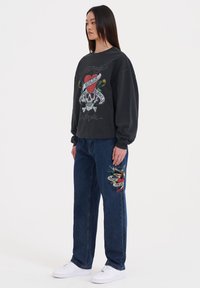 Czarna bluza z nadrukiem czaszki i serca, w połączeniu z niebieskimi dżinsami z haftowanymi detalami w postaci ptaka i wstęgi. Białe trampki.