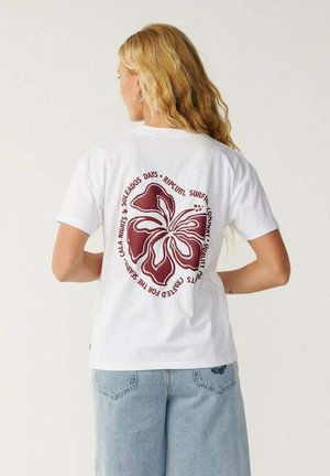 Femme blonde de dos portant un t-shirt blanc avec une grande fleur d'hibiscus rouge foncé et un texte, associée à un jean en denim bleu clair.
