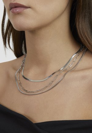 Three silver chains layered together, featuring a mix of flat and linked designs, against a bare neck, enhancing the sleek look.