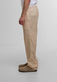 Lyse beige og kremfargede strippede bukser med elastisk midje. Materialet ser mykt ut; detalj med sidelomme synlig. Brukt med brune tresko.