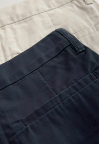 Deux pantalons étalés à plat : un beige et un bleu marine. Les deux présentent un tissu lisse avec des coutures visibles et des poches arrière.