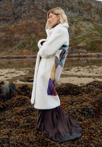 Lång, vit fuskpälsjacka med färggörande abstrakt mönster på sidan, bärs över en mörkbrun satindress. Slät textur och flödande form.