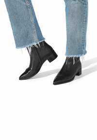 Botas de cuero negro hasta el tobillo con punta afilada y un tacón bajo cuadrado. Combinadas con jeans de mezclilla azul de dobladillo deshilachado.