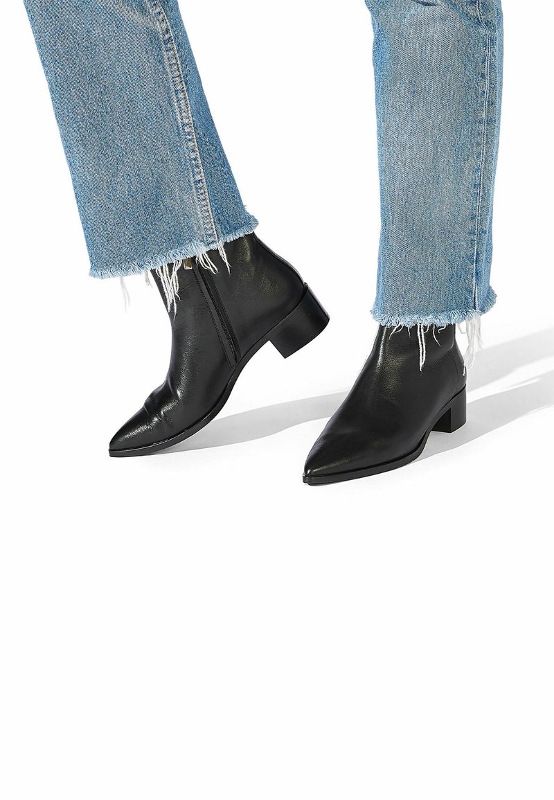 Botas de cuero negro hasta el tobillo con punta afilada y un tacón bajo cuadrado. Combinadas con jeans de mezclilla azul de dobladillo deshilachado.