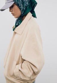 Beige bombardérka s voľným strihom, ktorá má golier, bočné vrecká a elastické manžety. Vzhľad dopĺňa zelený šál s paisley vzorom a čiapka.
