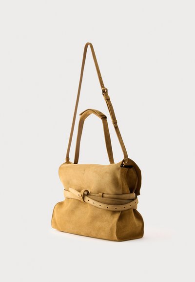 Sac à main en daim beige avec poignées supérieures, bandoulière réglable et ceinture décorative autour du milieu, sur un fond clair.