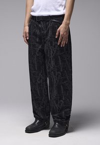 Μαύρο παντελόνι με υφή και αφηρημένο σχέδιο. Διαθέτει άνετη γραμμή, βασικές θηλιές για ζώνη και συνδυάζεται με μαύρα παπούτσια.