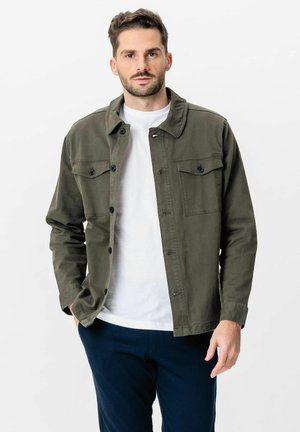 Mand iført en olivengrøn knapjakke, hvid T-shirt og mørkeblå bukser, stående med den ene hånd i lommen mod en hvid baggrund.