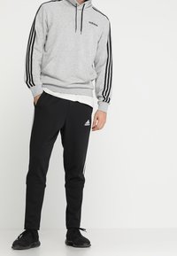 Felpa grigia con strisce nere, dotata di tasca a marsupio e cappuccio constringibile. Pantaloni sportivi neri con strisce bianche e logo sulla gamba.