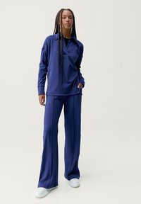 Námorně modrý sportovní set obsahující dlouhý rukáv s límcem a volné kalhoty se bočními švy, doplněné bílými teniskami.